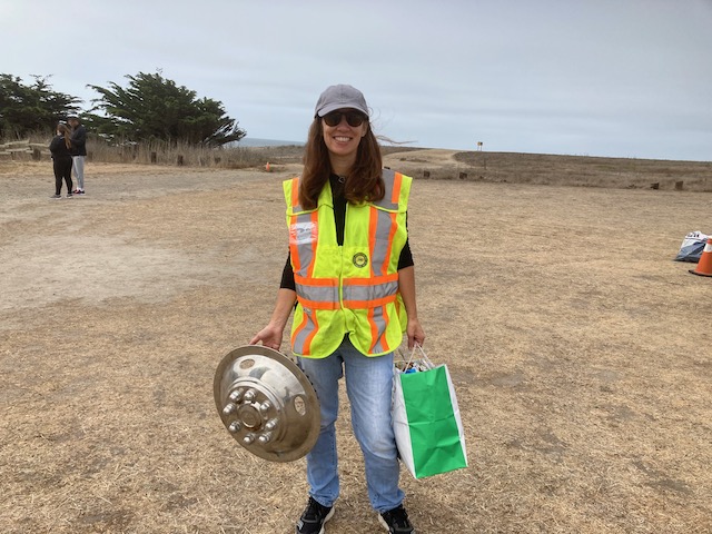 A woman in a high-vis vest at the beach cleanup, holding a hubcap and a shopping bag.