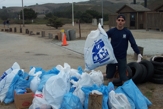 Person at cleanup day holding a bag of trash while surrounded by bags of trash.